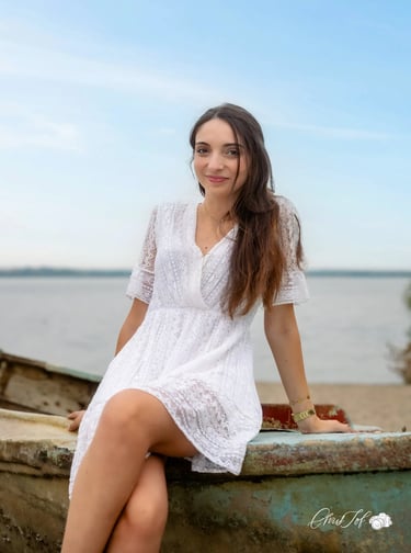 Jeune femme souriante en robe blanche en dentelle assise sur une barque au bord de l'eau à Canet 66.