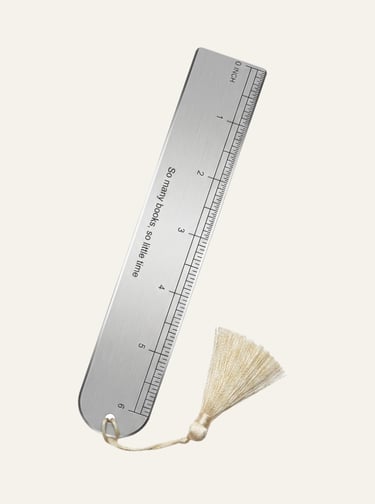 A brushed metal ruler bookmark with an engraved quote and a cream silk tassel on a plain background.