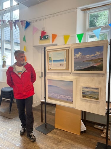 Artist Chris Gill standing beside framed coastal oil paintings at New Atlantic Wave Art Exhibition, 
