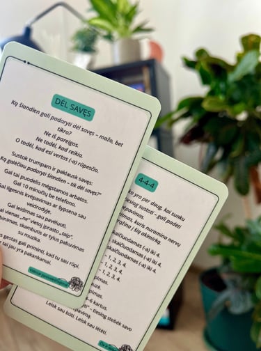 Emocinės gerovės kortelės laikomos rankoje, fone matosi kambarinės žalios gėlės