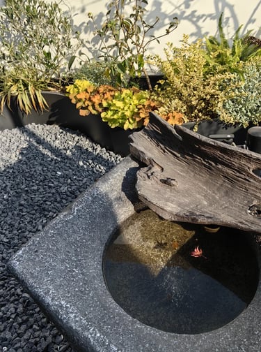 Modern outdoor water feature with natural stone, dark gravel, wood texture, and layered planting.