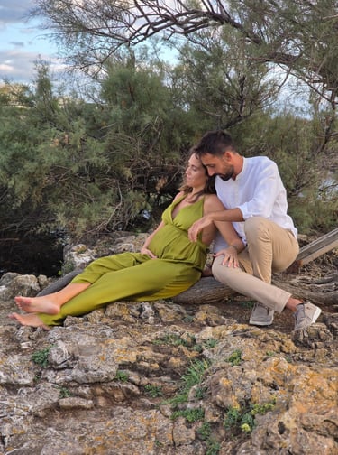 A family of three sitting on rocky terrain near a river with greenery in the background, enjoying an outdoor moment.