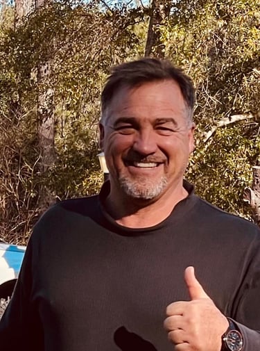 A smiling man with a goatee giving a thumbs up outdoors with trees and a truck in the background.