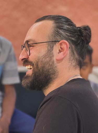 Smiling man with glasses, a beard, and a man bun hairstyle laughing during a social gathering.
