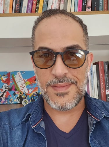 A man with a graying beard and glasses posing in front of a bookshelf with colorful books.