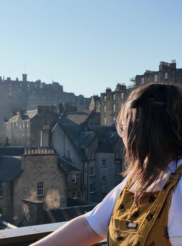 A women (Erin, owner of EB Socials) with glasses, brown hair and yellow dungagrees, looking out toward Edinburgh Castle