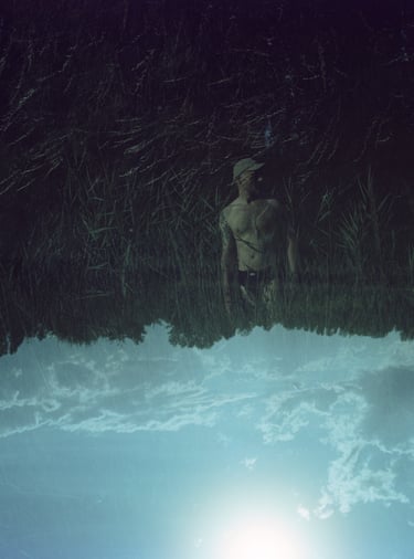 Surreal double exposure of a man in tall grass merged with an upside-down blue sky and clouds.