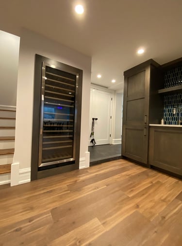 Modern home bar featuring a built-in wine cooler, dark cabinetry, and hardwood floors near a staircase.