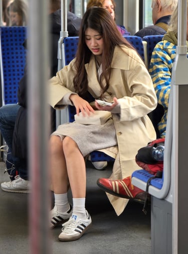 A young woman in a beige coat and white sneakers sits on a tram, looking down into her purse. Other passengers are nearby.