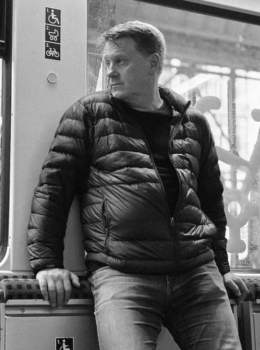A man in a puffy jacket and jeans leans on a train seat, gazing out the window. Accessibility signs are above. Black and whit