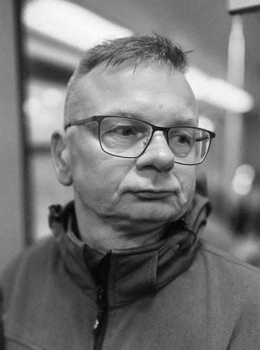 A middle-aged man with short hair and glasses looks sideways indoors, wearing a zip-up jacket. Black and white, blurred backg