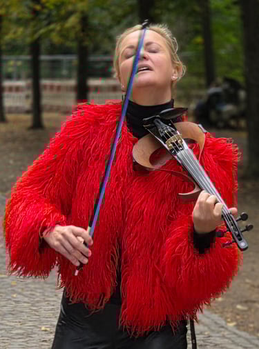 A woman in a bright red shaggy jacket plays her violin outdoors, eyes closed, head back, trees and cobblestones around her.