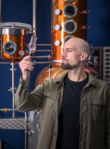 mastro distillatore che osserva del liquido in un bicchiere da degustazione davanti a un alambicco