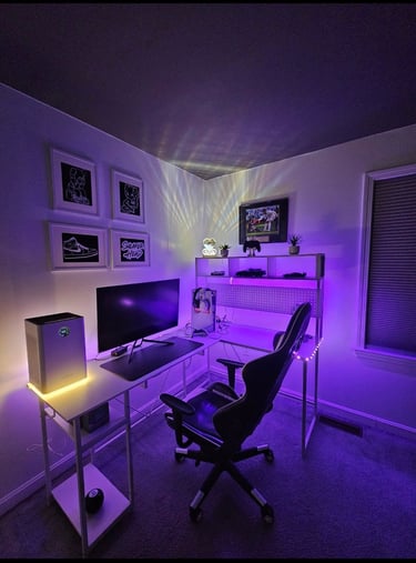 Modern gaming setup with an L-shaped desk, RGB lighting, and ergonomic chair in a purple-lit room.
