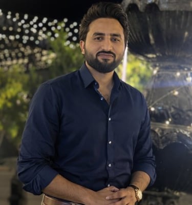 A bearded man in a navy blue button-down shirt posing outdoors at night with decorative lighting.