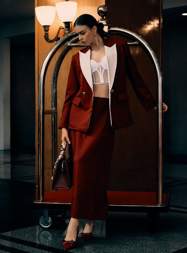 Model in a maroon blazer and maxi skirt holds a red handbag with a scarf, standing by a hotel lobby trolley.