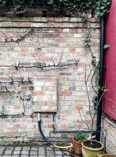 An electricity box attached to a garden brick wall painted in camouflage brick pattern.