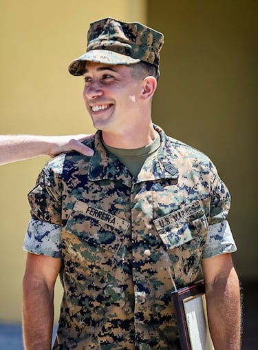 A smiling US Marine in MARPAT camouflage uniform holding a framed award certificate at a ceremony.