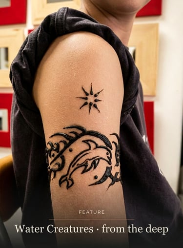 A person with a black henna arm tattoo featuring jumping dolphins, ocean waves, and a stylized sun.