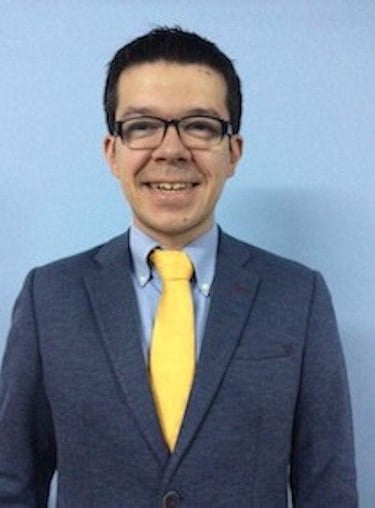 Smiling businessman in a blue suit and yellow tie wearing glasses against a blue background.
