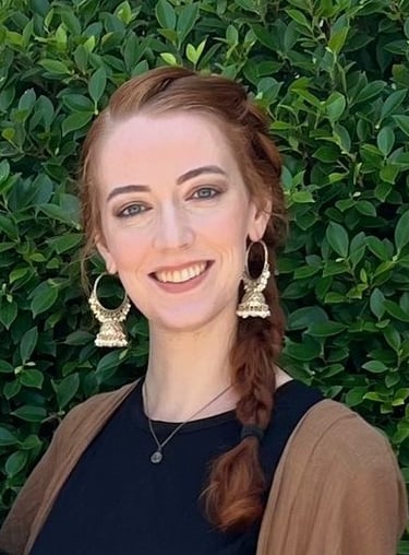 Smiling woman with red braided hair wearing gold Jhumka earrings against a green leaf hedge.