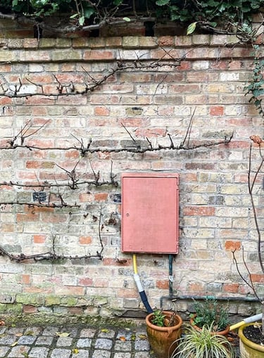 A bleached pink plastic electricity box attached to a garden brick wall.