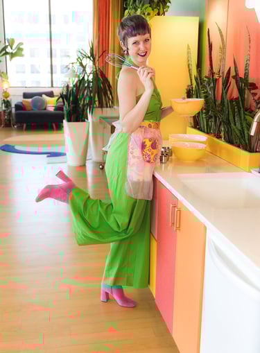 A woman in a green jumpsuit and pink boots baking in a colorful mid-century modern kitchen.