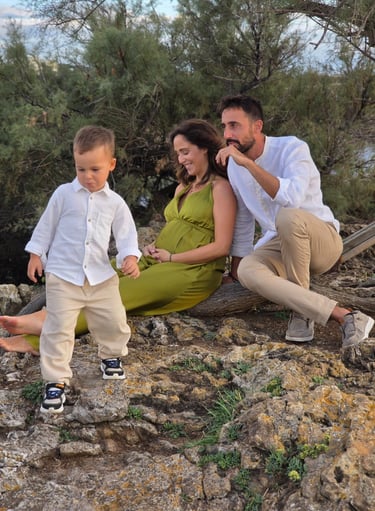 A family of three sitting on rocky terrain near a river with greenery in the background, enjoying an outdoor moment.