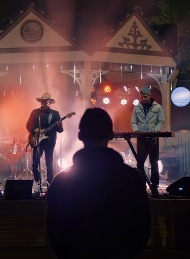 Live outdoor concert at night with a guitarist and keyboardist performing on a lit stage under a dec