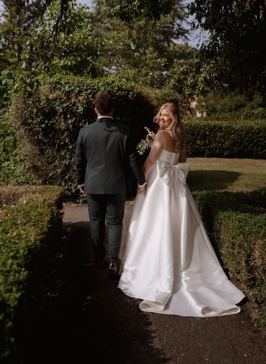 Smiling bride in a strapless white wedding dress with a large bow walking in a garden with her groom.