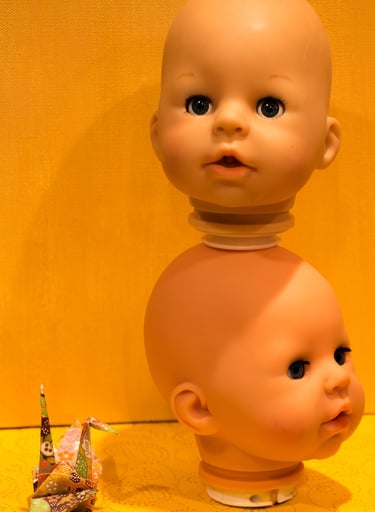 Small paper origami crane sitting next to 2 baby doll heads set on top of each other.