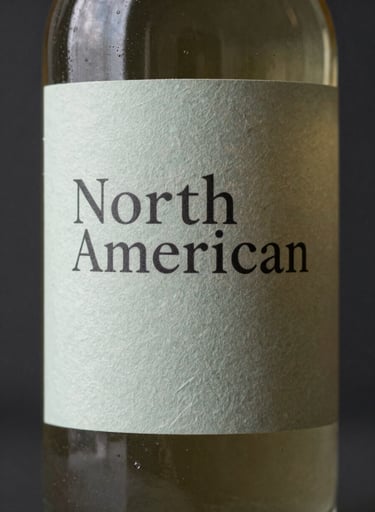 A close-up photograph of a premium glass bottle with a minimalist dusty sage paper label, featuring elegant typography, set against a dark charcoal background in a North American / US studio setting.