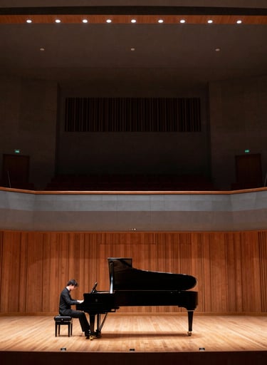 A wide-angle photography shot of a grand concert hall stage featuring a piano and a soloist's position. Warm, sophisticated lighting with dark gray and wood accents. North American / US cultural center.