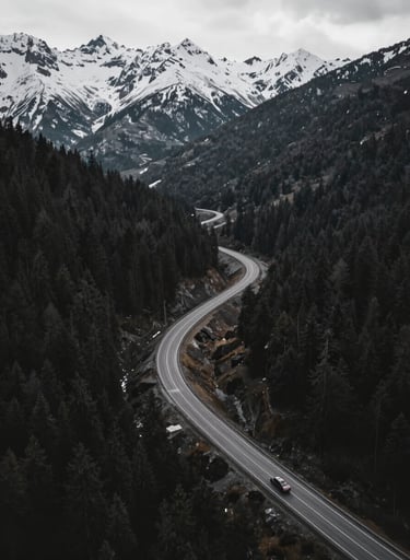 An ultra-wide 4K drone shot of a winding mountain road cutting through a dark forest. A single vehicle moves with speed, captured in a dynamic, sweeping motion. The contrast between the #1B263B shadows and the #E0E1DD snow-capped peaks is sharp.
