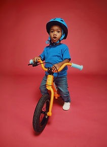A young boy wearing a blue helmet and shirt rides an orange balance bike against a red background.