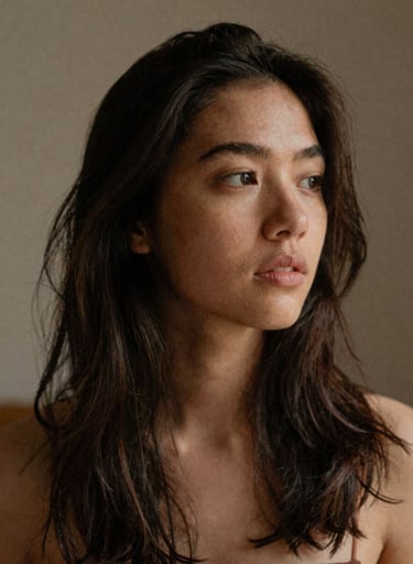 Candid, soft-focus portrait of a young woman with natural, slightly messy hair, looking away thoughtfully. Grainy film texture, underexposed, warm natural light, palette includes muted terracotta (#D1B499) and charcoal shadows (#3E352B).