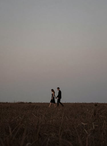 Wide landscape shot of a couple walking together in a field during blue hour. Artistic elegance with deep #20232B tones and soft #6E6D74 greys. The composition emphasizes the scale of the environment and the intimacy of the pair.