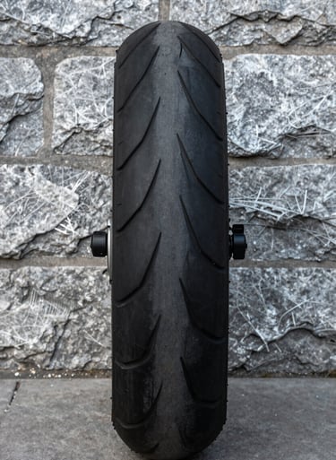 A minimalist detail of a jet black motorcycle tire against a weathered silver grey stone wall. The textures of the rubber and stone are emphasized by low-angle lighting. Global / Western urban environment.
