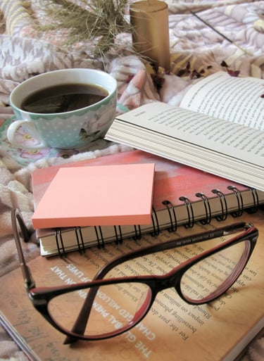 A pair of glases, a novel, a cup of tea, and a notebook sit on a shabby-chic tablecloth.