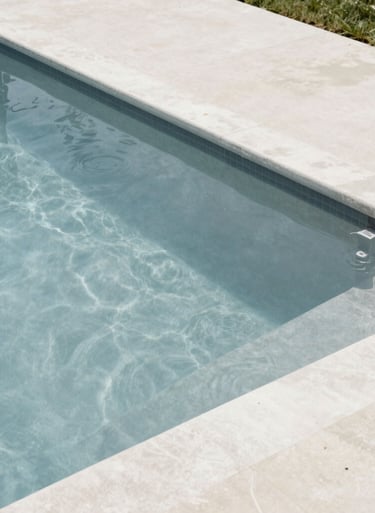 A high-angle shot of a classic North American suburban pool at midday. The water is a clear, soft blue-grey, contrasting with a clean off-white concrete deck. Minimalist, contemporary aesthetic.