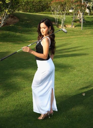 A stylish woman in a long white skirt holding a golf club on a lush green golf course.