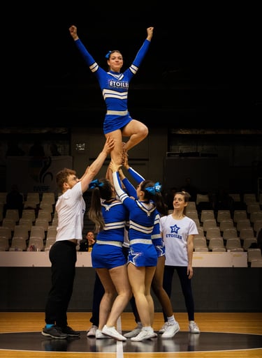 A cheerleading flyer in a blue uniform performs a stunt being lifted by her team during a competition.