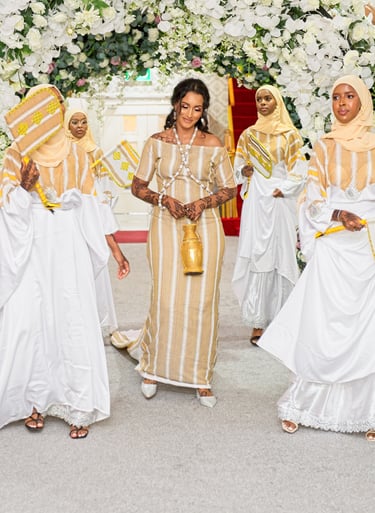 Happy Somalian bride and bridesmaids entering wedding venue all in traditional white & gold dresses, floral arch framing them