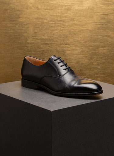 High-end product photography of a luxury leather shoe on a dark charcoal black pedestal. The background is brushed gold, with sharp lighting and clean lines. Modern and commercial style.