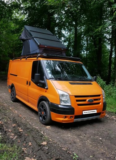 Orange Ford Transit camper van with a pop-top roof tent parked on a forest trail.