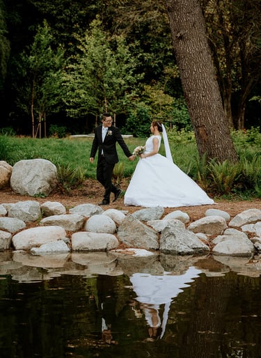 A newlywed couple walks hand-in-hand by a reflective pond during an outdoor garden wedding ceremony.