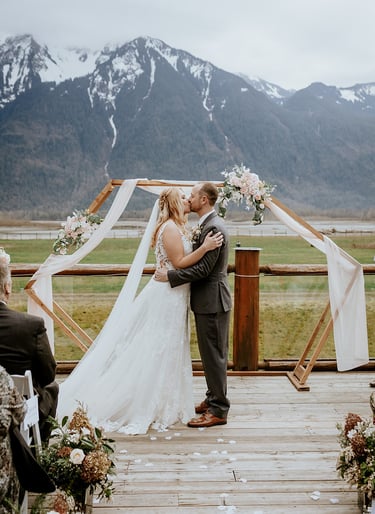 A bride and groom sharing their first kiss under a wooden geometric arch during an outdoor mountain wedding ceremony.