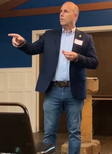 A standing man in jeans and a suit jacket pointing to the right the 