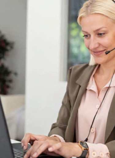 Blonde female customer support representative wearing a headset and working on a laptop in an office.