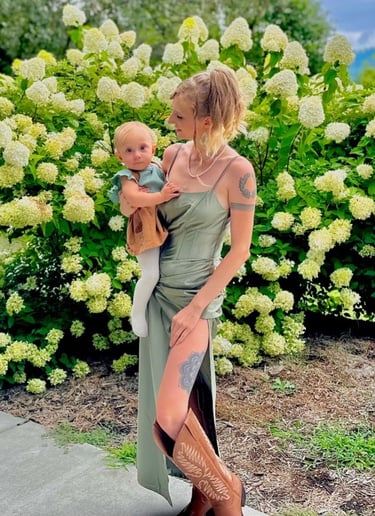 Jade in a green dress and boots holding baby Nova by flow bush, representing maternal instinct and caregiving